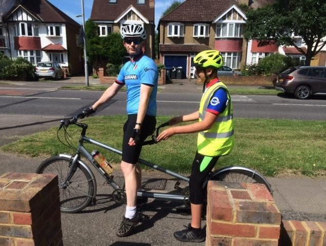 Image shows two male cyclists wearing bright cycling clothing, both sitting on a tandem bike. the front cyclist is sighted and the rear person is partially sighted. They are on a pavement with rows of houses in the background. 