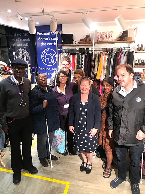 Image shows the KAB CEO Odette in the middle of a group of smiling visually impaired grow of member visitors. They are standing in the Vision Foundation charity shop, with a clothes rack behind them and a blue sign that says 'Fashion that doesn't cost the earth', with an illustration of a globe below the words.